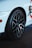 A close-up view of a car tire mounted on a glossy black rim. The body of the car is white with a sleek design, and a portion of a stylish decal is visible. The focus is on the tread and details of the wheel, highlighting its sporty features.