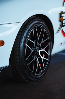 A close-up view of a car tire mounted on a glossy black rim. The body of the car is white with a sleek design, and a portion of a stylish decal is visible. The focus is on the tread and details of the wheel, highlighting its sporty features.