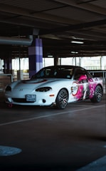 A white sports car is parked in a dimly lit parking garage. The side of the car features a vibrant anime-style illustration with bright pink accents. The car has a sporty look with black rims and red trim, and there's a distinctive pink tow hook at the front.