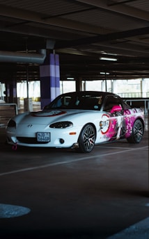 A white sports car is parked in a dimly lit parking garage. The side of the car features a vibrant anime-style illustration with bright pink accents. The car has a sporty look with black rims and red trim, and there's a distinctive pink tow hook at the front.