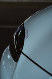 A close-up of a car's front section, focusing on the sleek design of the headlight and bodywork. The surface reflects dim lighting and there is a small decal with Japanese characters in purple.