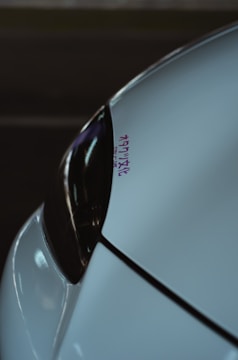 A close-up of a car's front section, focusing on the sleek design of the headlight and bodywork. The surface reflects dim lighting and there is a small decal with Japanese characters in purple.