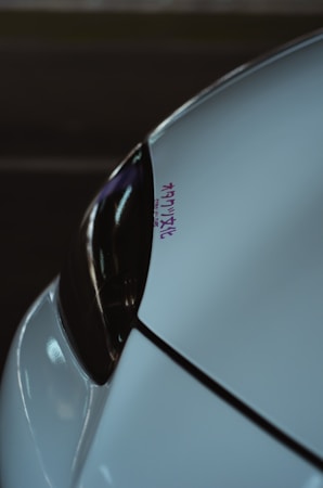 A close-up of a car's front section, focusing on the sleek design of the headlight and bodywork. The surface reflects dim lighting and there is a small decal with Japanese characters in purple.