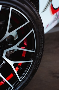 A close-up view of a car wheel with a stylish alloy rim. The wheel features a sleek black and silver spoke design with red accents visible through the spaces in the rim. The tire is branded with 'Hankook,' a well-known tire manufacturer.