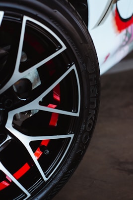 A close-up view of a car wheel with a stylish alloy rim. The wheel features a sleek black and silver spoke design with red accents visible through the spaces in the rim. The tire is branded with 'Hankook,' a well-known tire manufacturer.
