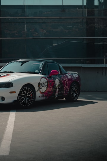 A stylishly customized car featuring anime-themed artwork on the side. The design includes a large graphic of a character with flowing hair. The car is parked in an urban setting, with a modern glass building facade in the background. The wheels have a unique spoke design, and the car's body has a glossy finish.