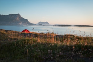 A scenic landscape featuring a vibrant red tent pitched on grassy terrain by a tranquil body of water, with distant mountains under a clear blue sky. The sunlight creates a warm glow, highlighting the natural beauty of the surroundings.