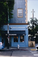 A blue building corner with 'Boulangerie' written on it, featuring a black awning above the entrance. A single window is visible above the sign. There are trees on either side, a fire hydrant on the sidewalk, and a blue car parked nearby.