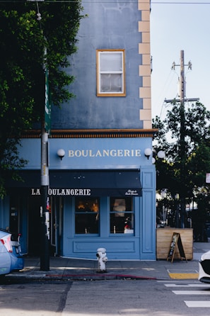 A blue building corner with 'Boulangerie' written on it, featuring a black awning above the entrance. A single window is visible above the sign. There are trees on either side, a fire hydrant on the sidewalk, and a blue car parked nearby.