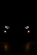 A pair of car headlights and fog lights are illuminated in a dark, night-time setting. The lights are brightly shining against a pitch-black background, giving the impression of a vehicle approaching or standing still in darkness. The headlights have a modern, angular design, and there are two smaller, round fog lights positioned below them.