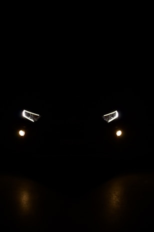 A pair of car headlights and fog lights are illuminated in a dark, night-time setting. The lights are brightly shining against a pitch-black background, giving the impression of a vehicle approaching or standing still in darkness. The headlights have a modern, angular design, and there are two smaller, round fog lights positioned below them.
