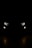 A pair of car headlights and fog lights are illuminated in a dark, night-time setting. The lights are brightly shining against a pitch-black background, giving the impression of a vehicle approaching or standing still in darkness. The headlights have a modern, angular design, and there are two smaller, round fog lights positioned below them.
