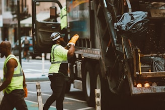 Workers are performing garbage collection on a city street. Two individuals wearing high-visibility safety vests and work gloves are seen near a garbage truck. One worker is placing a trash bag into the back of the truck while the other stands nearby.