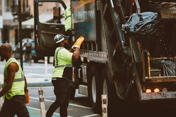 Workers are performing garbage collection on a city street. Two individuals wearing high-visibility safety vests and work gloves are seen near a garbage truck. One worker is placing a trash bag into the back of the truck while the other stands nearby.