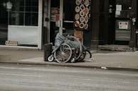 A folded wheelchair covered with a grey cloth is placed on a sidewalk in an urban setting. There are storefronts in the background, including one with a 'We’re Open' sign and a menu board displaying food images.