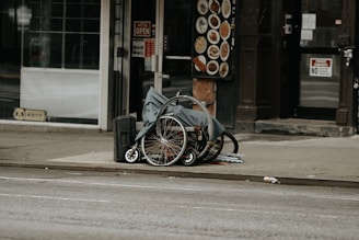 A folded wheelchair covered with a grey cloth is placed on a sidewalk in an urban setting. There are storefronts in the background, including one with a 'We’re Open' sign and a menu board displaying food images.