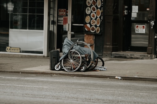 A folded wheelchair covered with a grey cloth is placed on a sidewalk in an urban setting. There are storefronts in the background, including one with a 'We’re Open' sign and a menu board displaying food images.