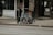 A folded wheelchair covered with a grey cloth is placed on a sidewalk in an urban setting. There are storefronts in the background, including one with a 'We’re Open' sign and a menu board displaying food images.
