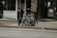 A folded wheelchair covered with a grey cloth is placed on a sidewalk in an urban setting. There are storefronts in the background, including one with a 'We’re Open' sign and a menu board displaying food images.