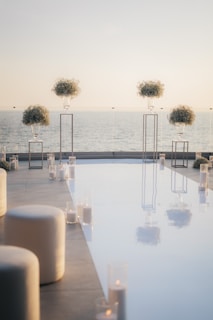 A beautifully arranged outdoor setting with a reflective pool surrounded by tall glass candle holders. Elegant white seating and elevated floral arrangements stand against a serene ocean backdrop under a clear sky.