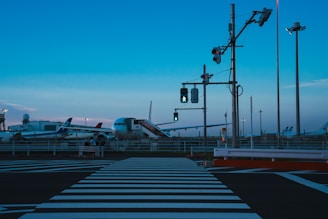 A twilight scene at an airport features several airplanes parked near the tarmac. A prominently visible zebra crossing leads to a traffic signal with illuminated pedestrian signs. The sky has a gradient of blue hues, and tall poles and lighting towers are scattered across the area.