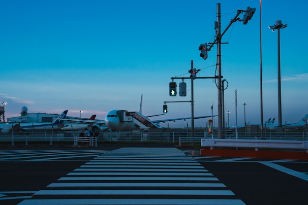 A twilight scene at an airport features several airplanes parked near the tarmac. A prominently visible zebra crossing leads to a traffic signal with illuminated pedestrian signs. The sky has a gradient of blue hues, and tall poles and lighting towers are scattered across the area.