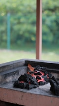 A pile of black charcoal is glowing with red embers in an outdoor grill. The charcoal pieces are arranged in a pile, indicating they might be ready for cooking. In the background, there is a blurred view of greenery, possibly a garden, with soft lighting suggesting a calm day.