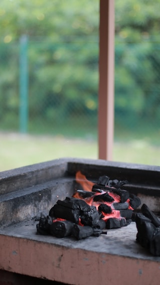 A pile of black charcoal is glowing with red embers in an outdoor grill. The charcoal pieces are arranged in a pile, indicating they might be ready for cooking. In the background, there is a blurred view of greenery, possibly a garden, with soft lighting suggesting a calm day.