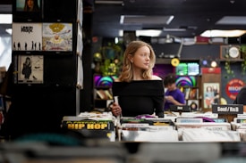 A woman stands in a record store, browsing through vinyl records. She is wearing an off-the-shoulder black top and has wavy blonde hair. The store is filled with various music albums, categorized under genres like Hip Hop, Jazz, and Soul/R&B. There are music posters and decorations on the walls, adding a retro vibe to the scene.