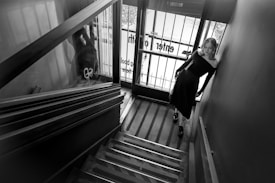 A black and white photograph of a woman in an off-the-shoulder dress standing at the bottom of a staircase. She leans against the wall near a glass door with bars, through which outside signage is partially visible. The stairwell features reflective surfaces and a slightly angular perspective, creating a modern and slightly dramatic scene.