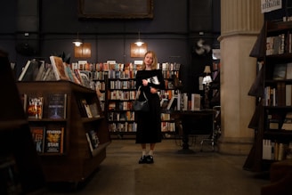 A person stands in a dimly lit bookstore, surrounded by shelves filled with books. The interior features warm lighting and wooden furnishings, creating a cozy atmosphere. The individual holds a book and appears to be contemplative.