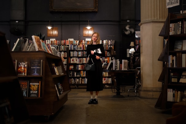 A person stands in a dimly lit bookstore, surrounded by shelves filled with books. The interior features warm lighting and wooden furnishings, creating a cozy atmosphere. The individual holds a book and appears to be contemplative.