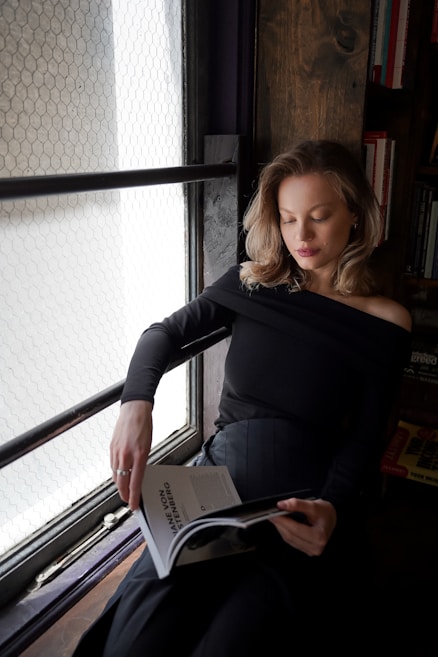 A person with blonde hair is seated by a window, reading a magazine. The setting is cozy and serene, with books on a shelf visible in the background. The person is wearing a black, off-the-shoulder top, and is focused on the magazine, which appears to be on fashion or lifestyle.