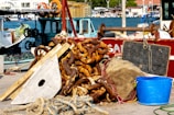 A pile of rusty chains and marine equipment lie on a dock next to several boats. The items include a large piece of wood, rope, and a blue plastic bucket. In the background, the ocean and harbor with additional boats can be seen, indicating an active fishing or maritime setting.