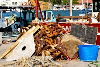 A pile of rusty chains and marine equipment lie on a dock next to several boats. The items include a large piece of wood, rope, and a blue plastic bucket. In the background, the ocean and harbor with additional boats can be seen, indicating an active fishing or maritime setting.
