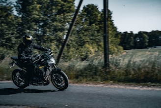 A motorcyclist dressed in black gear rides a sleek, dark motorcycle on a narrow road. The road is bordered by tall grass and dense, leafy trees under a bright sky. The motorcyclist appears to be riding at speed, conveying a sense of motion and freedom.