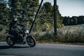 A motorcyclist dressed in black gear rides a sleek, dark motorcycle on a narrow road. The road is bordered by tall grass and dense, leafy trees under a bright sky. The motorcyclist appears to be riding at speed, conveying a sense of motion and freedom.