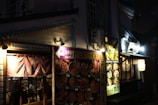 A dimly lit street scene featuring traditional Japanese architecture with illuminated signs and banners. The storefronts display a variety of colorful food images, suggesting a focus on seafood cuisine. There are paper lanterns and wooden accents, creating a cozy and inviting atmosphere.