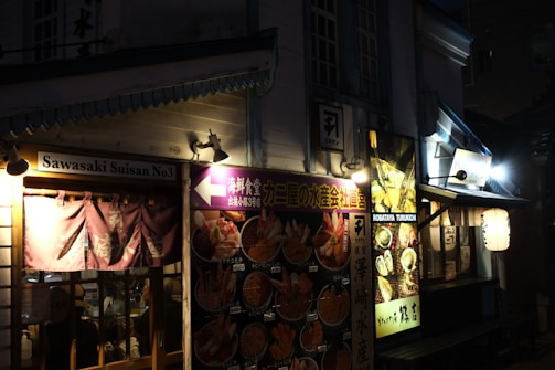 A dimly lit street scene featuring traditional Japanese architecture with illuminated signs and banners. The storefronts display a variety of colorful food images, suggesting a focus on seafood cuisine. There are paper lanterns and wooden accents, creating a cozy and inviting atmosphere.