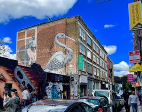 A vibrant urban street scene with several pieces of mural art on the side of a building. A large bird mural spans the height of the brick wall, while a skull artwork is visible on a smaller building. Various signs, including those for a vegan cafe and a retail unit, can be seen. People are walking and standing around, and several parked cars line the street.