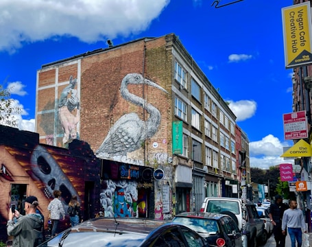 A vibrant urban street scene with several pieces of mural art on the side of a building. A large bird mural spans the height of the brick wall, while a skull artwork is visible on a smaller building. Various signs, including those for a vegan cafe and a retail unit, can be seen. People are walking and standing around, and several parked cars line the street.