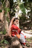 A young child dressed in traditional attire sits on a tree stump in a lush, green outdoor setting. The child wears a red and white outfit with a shawl draped over the head and is holding a tablet or similar device. Leaning on one hand with a thoughtful expression, the child is surrounded by greenery, including large leaves and trailing plants.