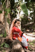 A young child dressed in traditional attire sits on a tree stump in a lush, green outdoor setting. The child wears a red and white outfit with a shawl draped over the head and is holding a tablet or similar device. Leaning on one hand with a thoughtful expression, the child is surrounded by greenery, including large leaves and trailing plants.