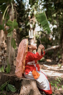 A child wearing traditional attire sits on a tree stump in a lush forest. The child is dressed in a bright red outfit with gold detailing and a pink headscarf. They hold a small object, possibly a flute, close to their face while surrounded by greenery.