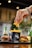 A hand adorned with a bracelet is placing a few French fries into a black container labeled with a fist graphic and 'Smashed is Better' text. The container is on a table alongside two hamburgers and three green glass bottles with colorful labels, likely containing beverages. The background is softly blurred, suggesting an outdoor setting.