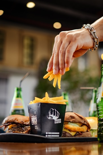 A hand adorned with a bracelet is placing a few French fries into a black container labeled with a fist graphic and 'Smashed is Better' text. The container is on a table alongside two hamburgers and three green glass bottles with colorful labels, likely containing beverages. The background is softly blurred, suggesting an outdoor setting.