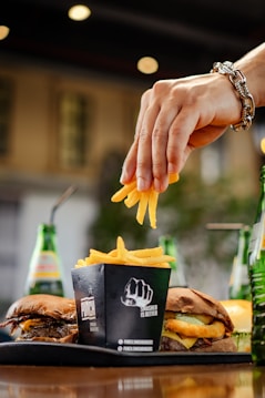 A hand adorned with a bracelet is placing a few French fries into a black container labeled with a fist graphic and 