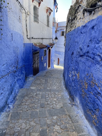 A narrow cobblestone alleyway flanked by vibrant blue walls, characteristic of traditional Moroccan architecture. The buildings feature iron window grilles and irregular stonework. Sunlight brightens the pathway, and a street lamp hangs from one side of the alley.