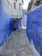 A narrow cobblestone alleyway flanked by vibrant blue walls, characteristic of traditional Moroccan architecture. The buildings feature iron window grilles and irregular stonework. Sunlight brightens the pathway, and a street lamp hangs from one side of the alley.