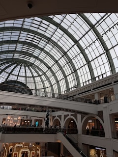 An interior of a shopping mall with a grand, arched glass ceiling that allows natural light to fill the space. The architecture includes ornate railings and multiple levels with escalators connecting the floors. Several luxury retail stores can be seen on the lower level.